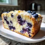 Close-up of a moist slice of blueberry bread, packed with berries and topped with white glaze.