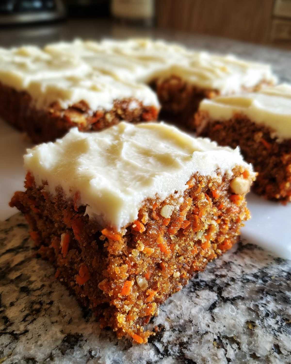 A close-up of a thick, moist square of carrot cake bars topped with cream cheese frosting, showing shredded carrots and nuts inside.