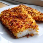 Close-up of two golden-brown, crispy parmesan crusted tilapia fillets on a white plate.