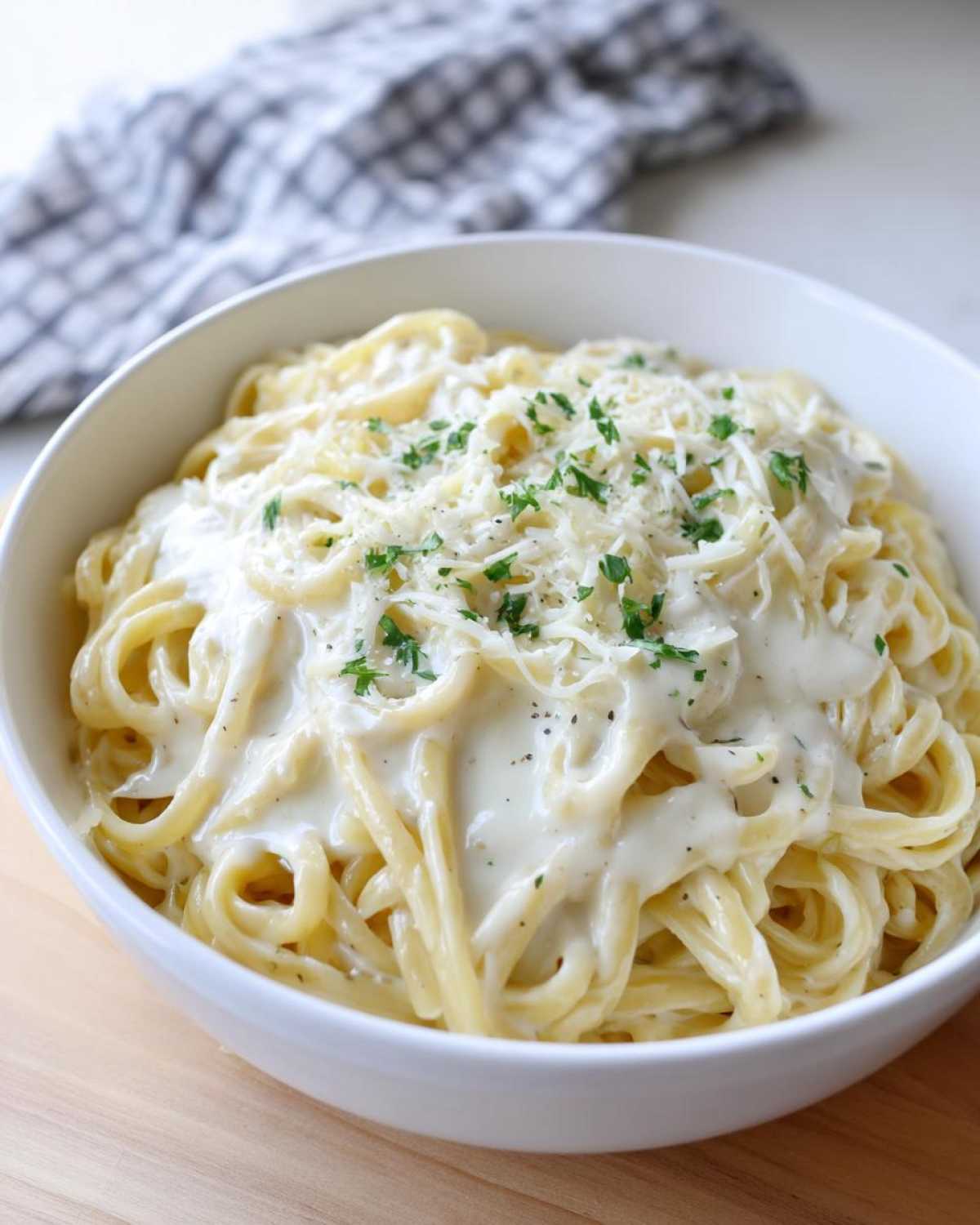 A white bowl filled with spaghetti generously coated in a rich cream sauce, topped with grated cheese and parsley.