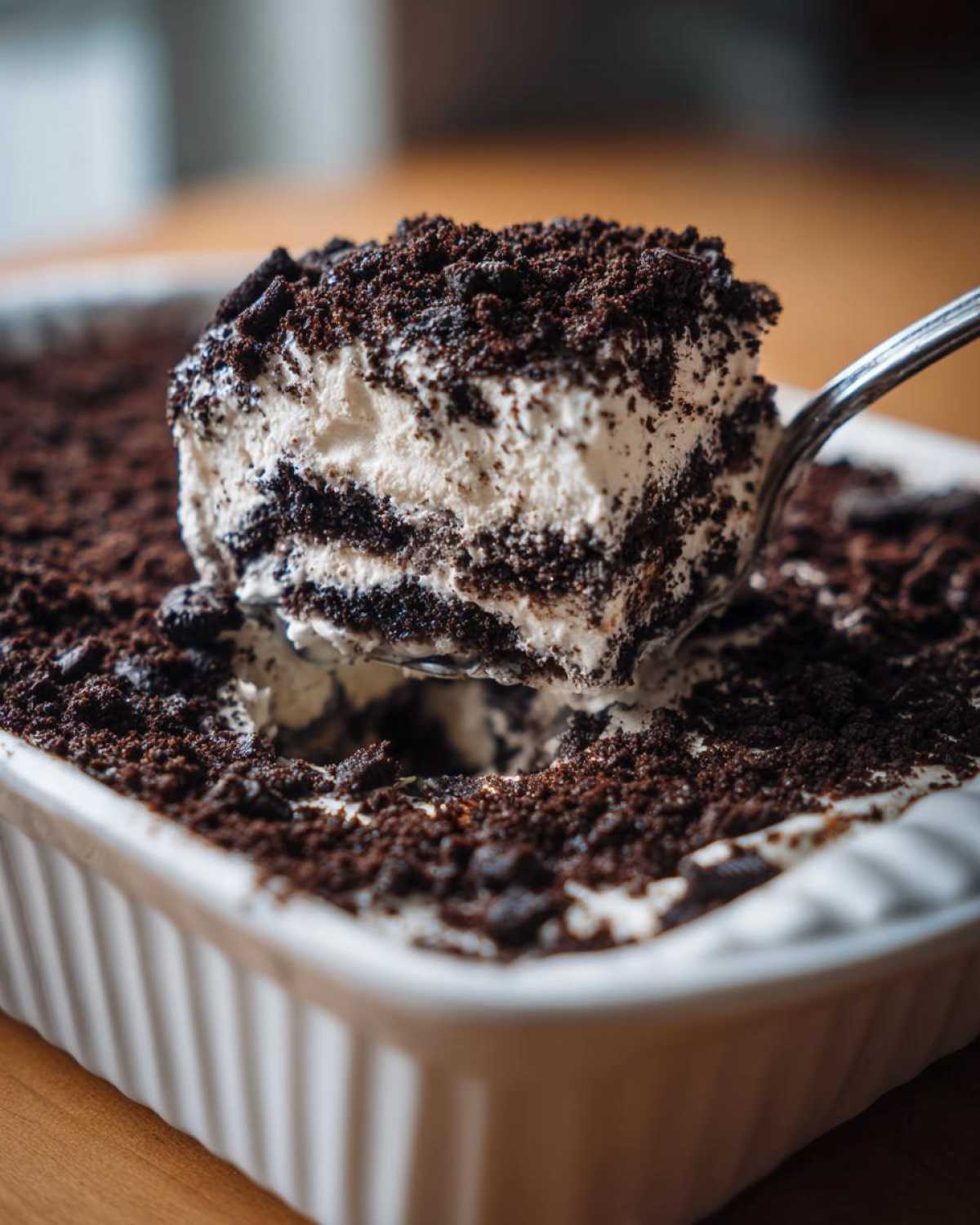 A spoonful of creamy, layered dirt cake being lifted from a white baking dish covered in chocolate crumbs.