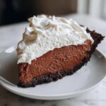 A perfect slice of chocolate cream pie featuring a dark crust, rich chocolate filling, and thick whipped cream topping.
