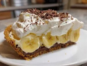 A decadent slice of banoffee pie featuring a graham cracker crust, chocolate layer, sliced bananas, and whipped cream topped with chocolate shavings.