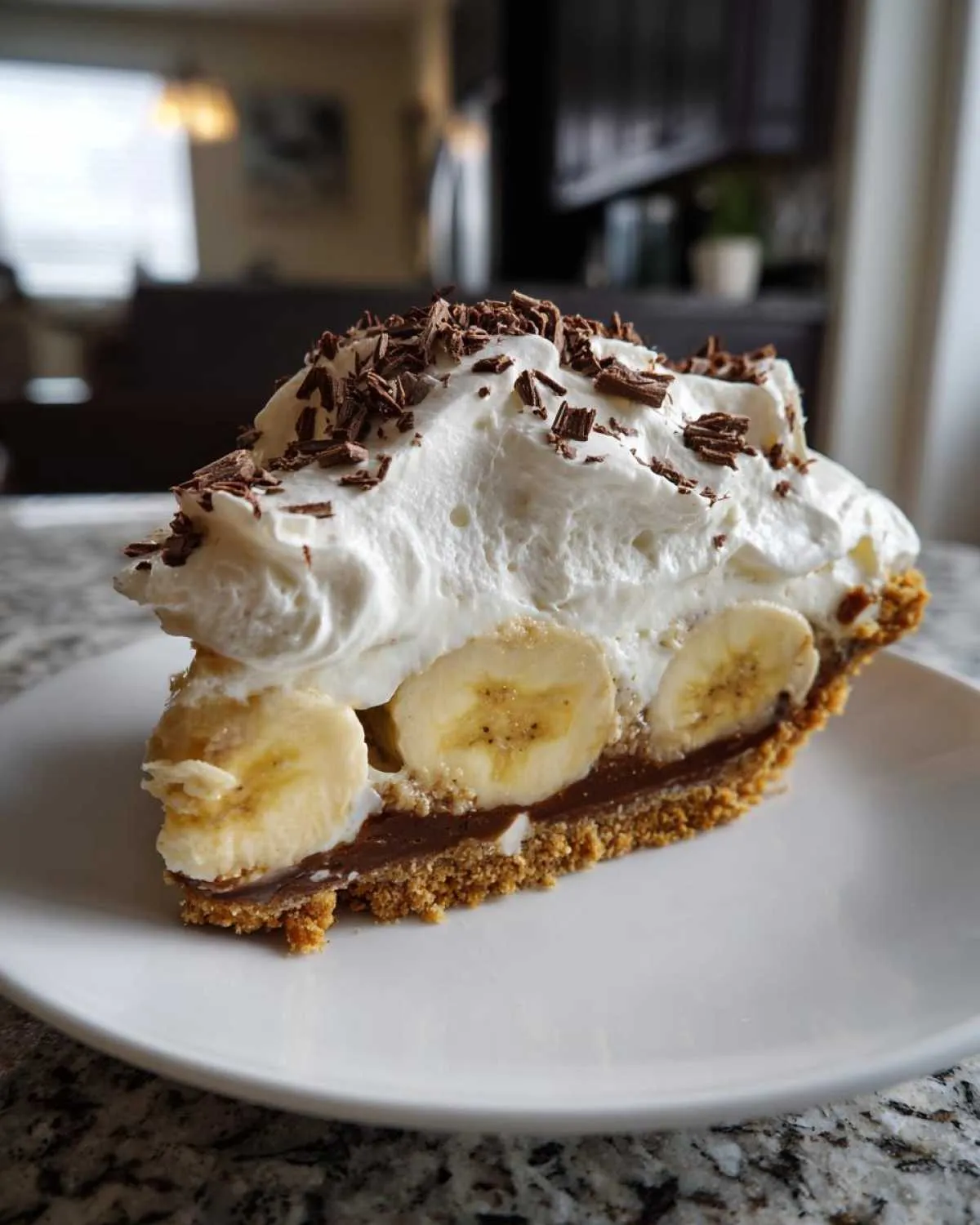 A perfect slice of banoffee pie featuring a graham cracker crust, chocolate filling, banana slices, and whipped cream topped with chocolate shavings.