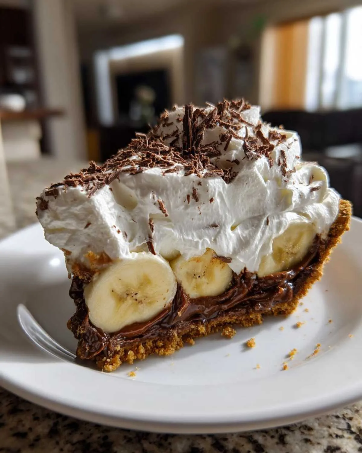A generous slice of banoffee pie featuring a graham cracker crust, bananas, chocolate filling, and whipped cream topped with chocolate shavings.