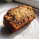 Close-up of a moist slice of sugar free banana bread with visible fruit/raisins, sitting on a light plate.