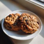 Two soft oatmeal raisin cookies resting on a small white plate, illuminated by bright sunlight.
