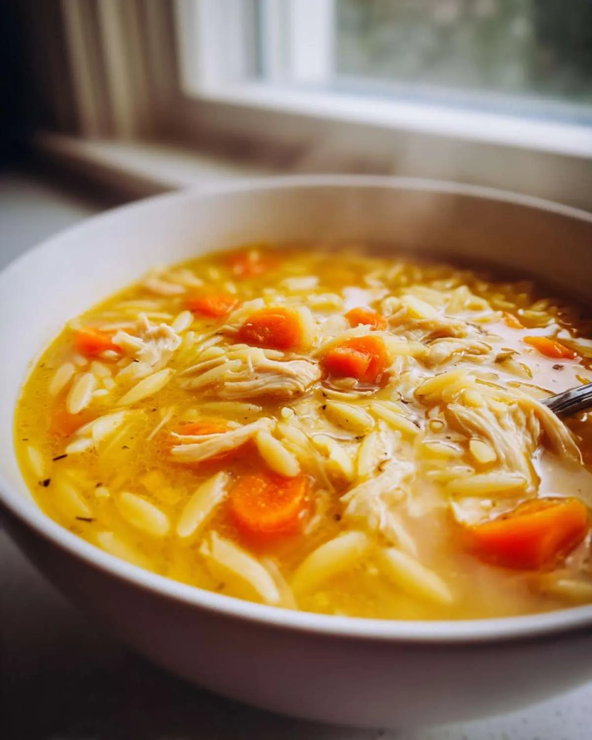 Close-up of a steaming white bowl filled with rich, yellow chicken orzo soup featuring shredded chicken and bright orange carrots.