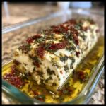 A log of creamy marinated cheese covered in herbs, garlic, and sun-dried tomatoes, submerged in olive oil.
