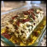 A log of creamy marinated cheese covered in herbs, garlic, and sun-dried tomatoes, submerged in olive oil.