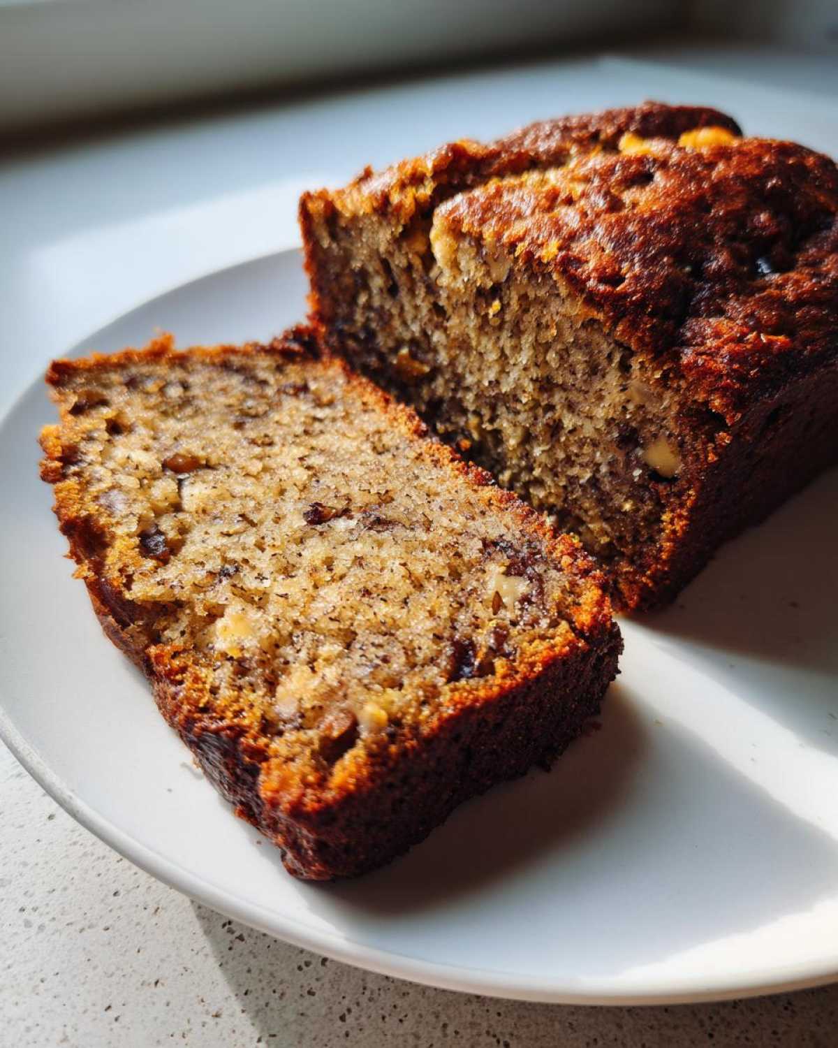 Two slices of moist sugar free banana bread, showing nuts and dark crumb texture, served on a white plate.
