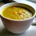 A close-up shot of thick, green pea soup with visible chunks of carrot served in a white mug.