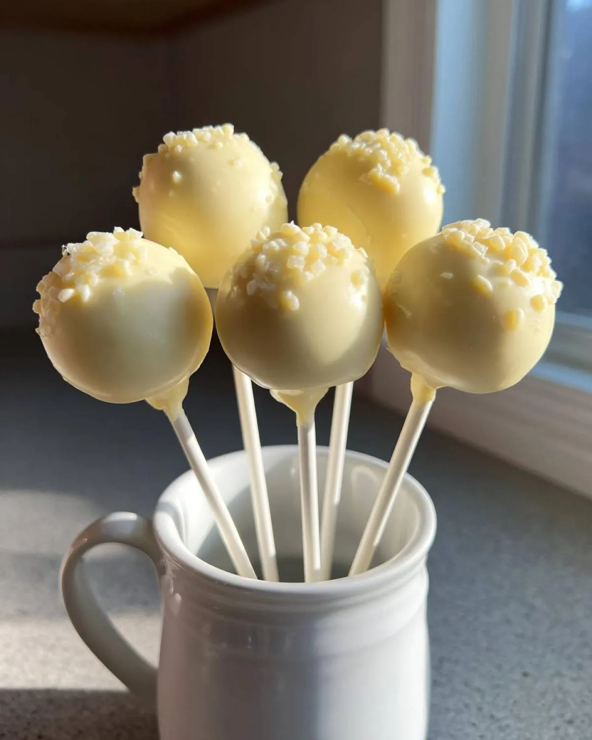 Five white chocolate cake pops with crunchy white sprinkles standing upright in a white ceramic mug.