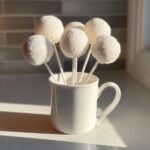 A bouquet of white, round cake pops covered in white sprinkles, standing upright in a white mug.