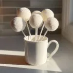 A bouquet of white, round cake pops covered in white sprinkles, standing upright in a white mug.