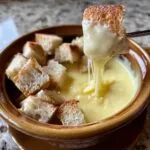 A piece of toasted bread being dipped into a pot of creamy, melted cheese fondue recipe, showing long cheese pulls.