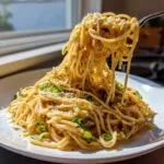 A fork lifts a large portion of saucy garlic noodles, topped with Parmesan and green onions, from a white plate.