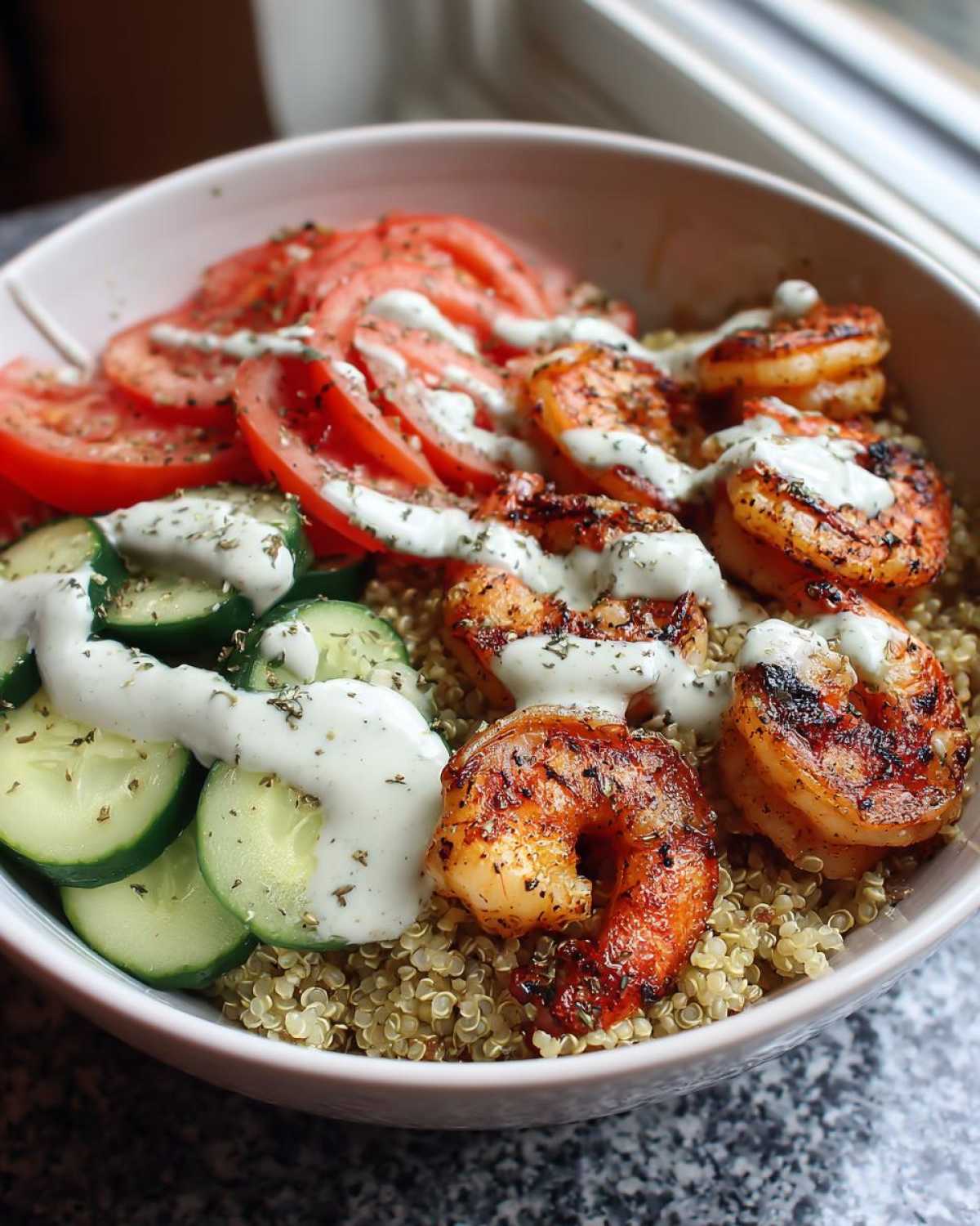 A close-up of a healthy grilled shrimp bowl featuring seasoned shrimp, quinoa, sliced cucumbers, tomatoes, and a creamy white sauce.