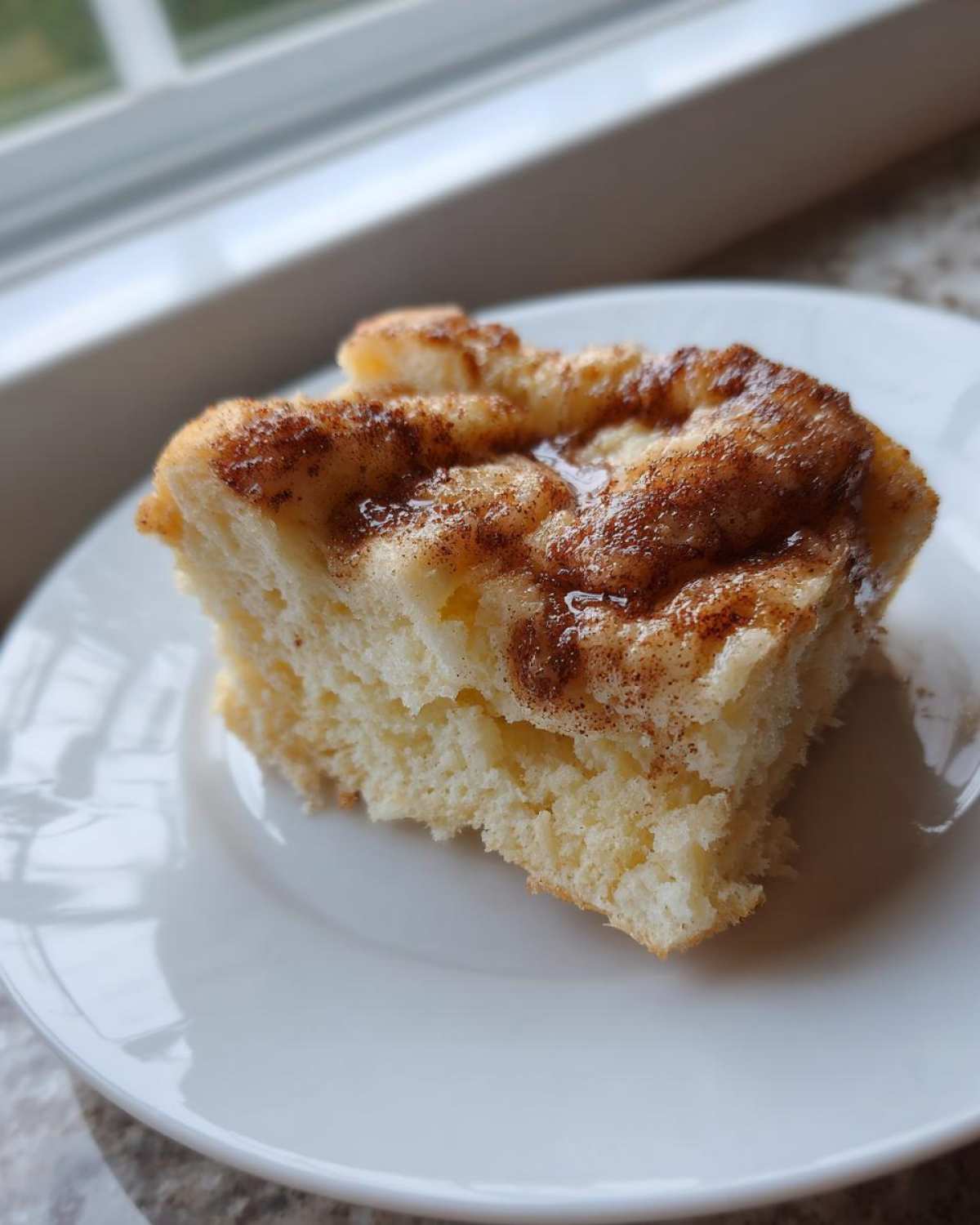 A close-up of a soft, fluffy square slice of amish cinnamon flop topped with a brown sugar and cinnamon mixture, served on a white plate.