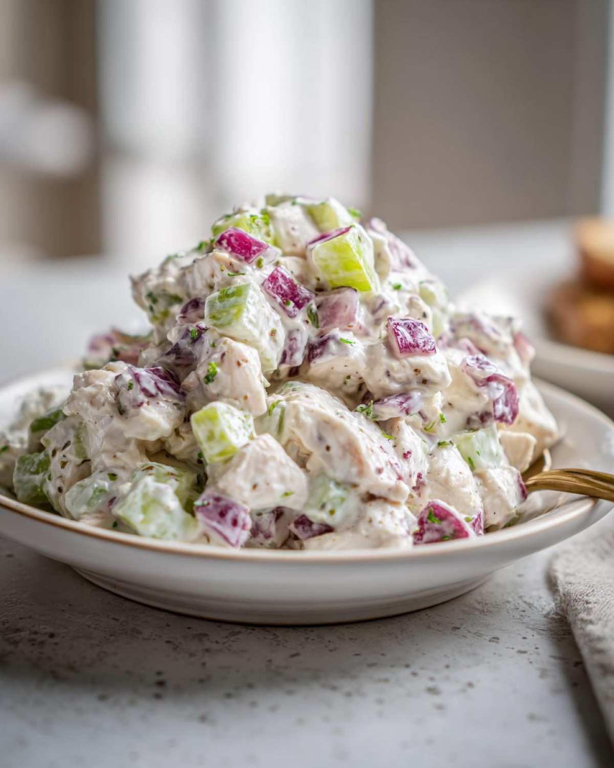 A close-up of creamy baked marinated chicken salad mixed with diced red onion and celery in a white bowl.