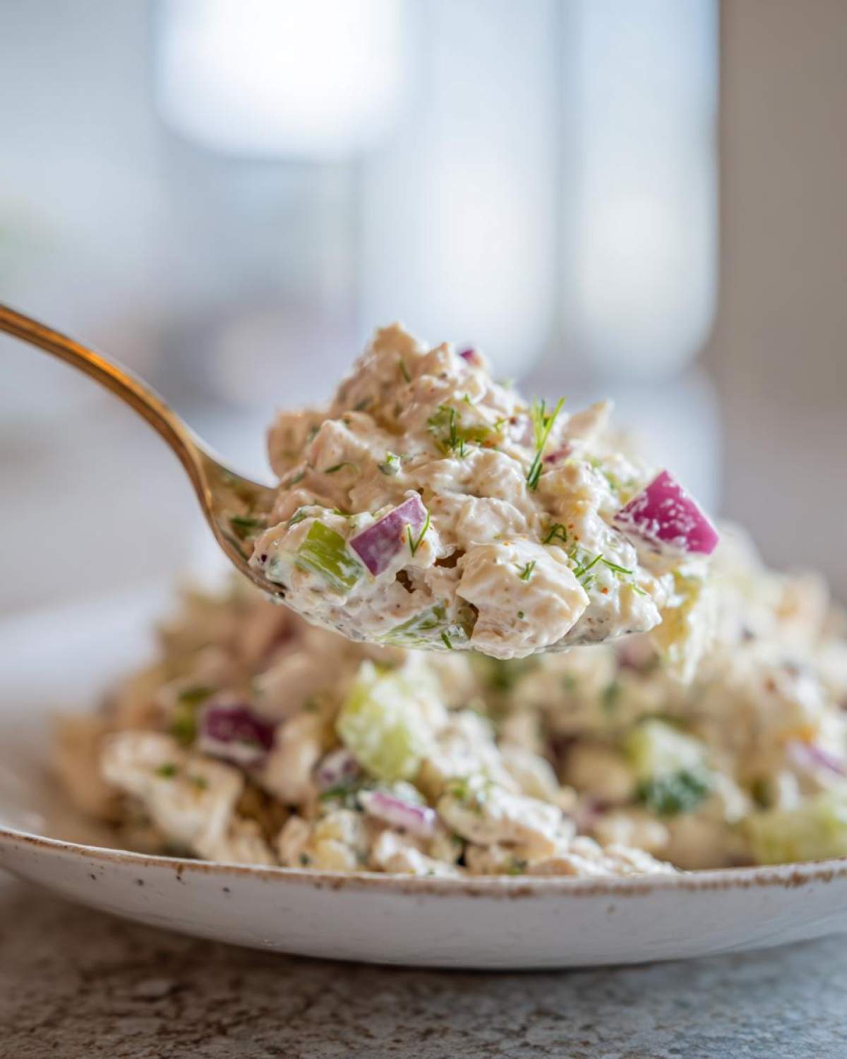 A spoonful of creamy baked marinated chicken salad featuring chunks of chicken, red onion, and celery, lifted above a bowl.