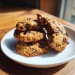A stack of four moist banana bread chocolate chip cookies on a white plate, one broken open.