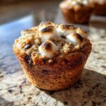 A close-up of a freshly baked banana s'mores muffin topped with toasted marshmallows and chocolate chips.