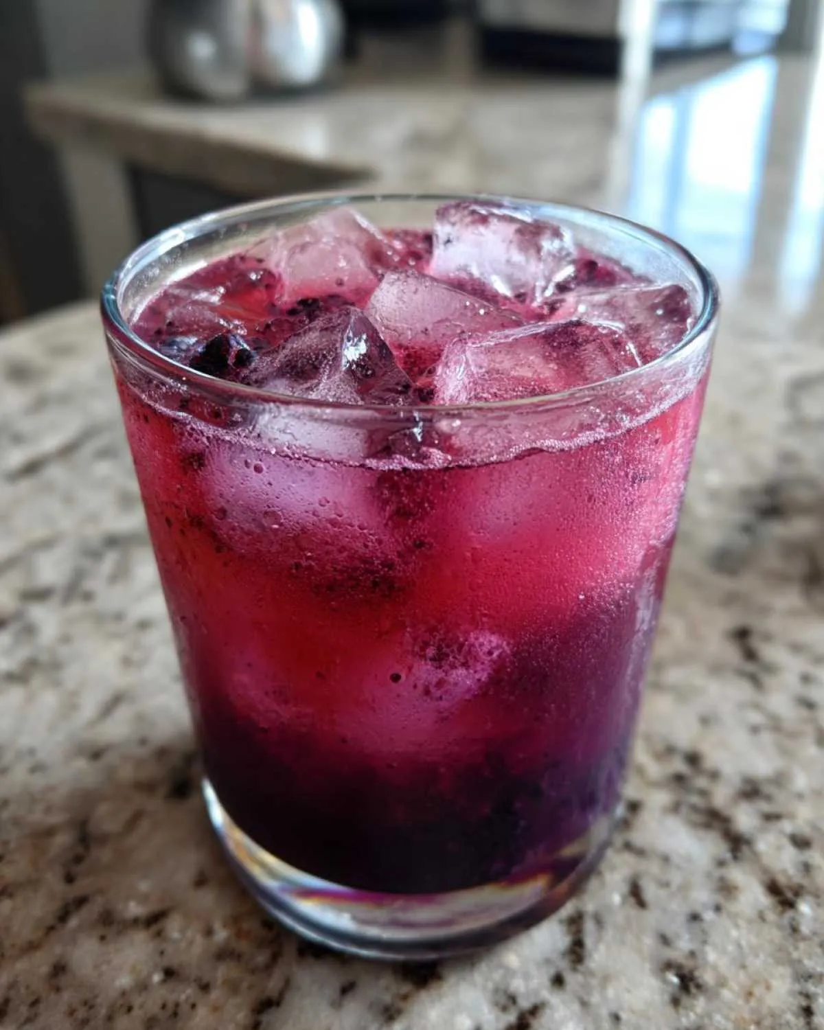 Close-up of a vibrant, icy blackberry coconut pink drink in a glass on a granite countertop.