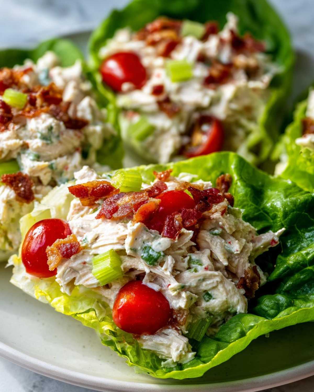 Close-up of blt chicken salad served in crisp lettuce cups, topped with bacon and cherry tomatoes.
