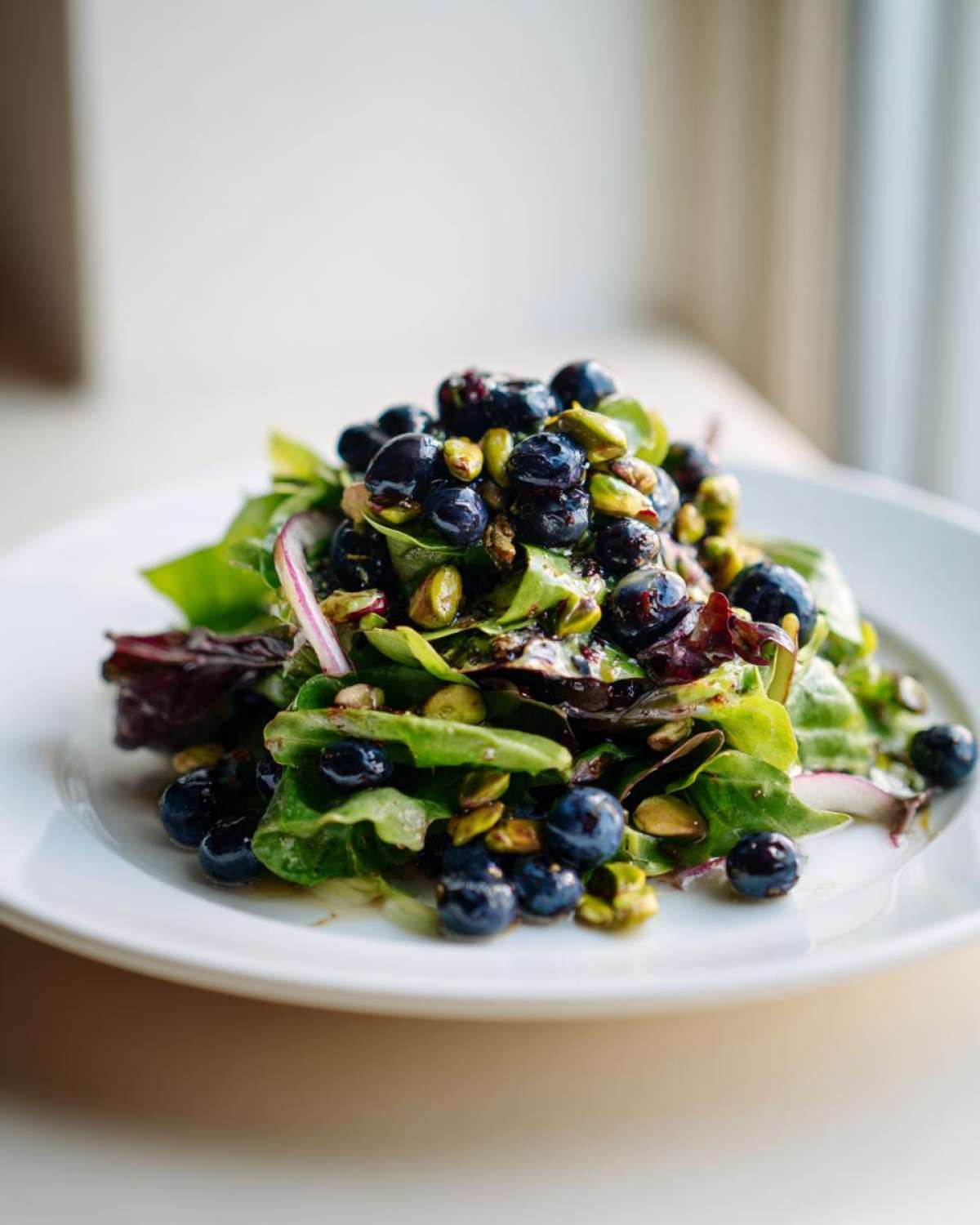 A vibrant mound of blueberry pistachio spring salad featuring mixed greens, fresh blueberries, and green pistachios on a white plate.