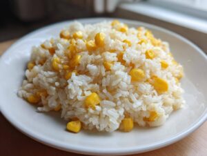 A close-up of freshly made buttered corn rice served on a white plate, highlighting the fluffy white rice and bright yellow corn kernels.