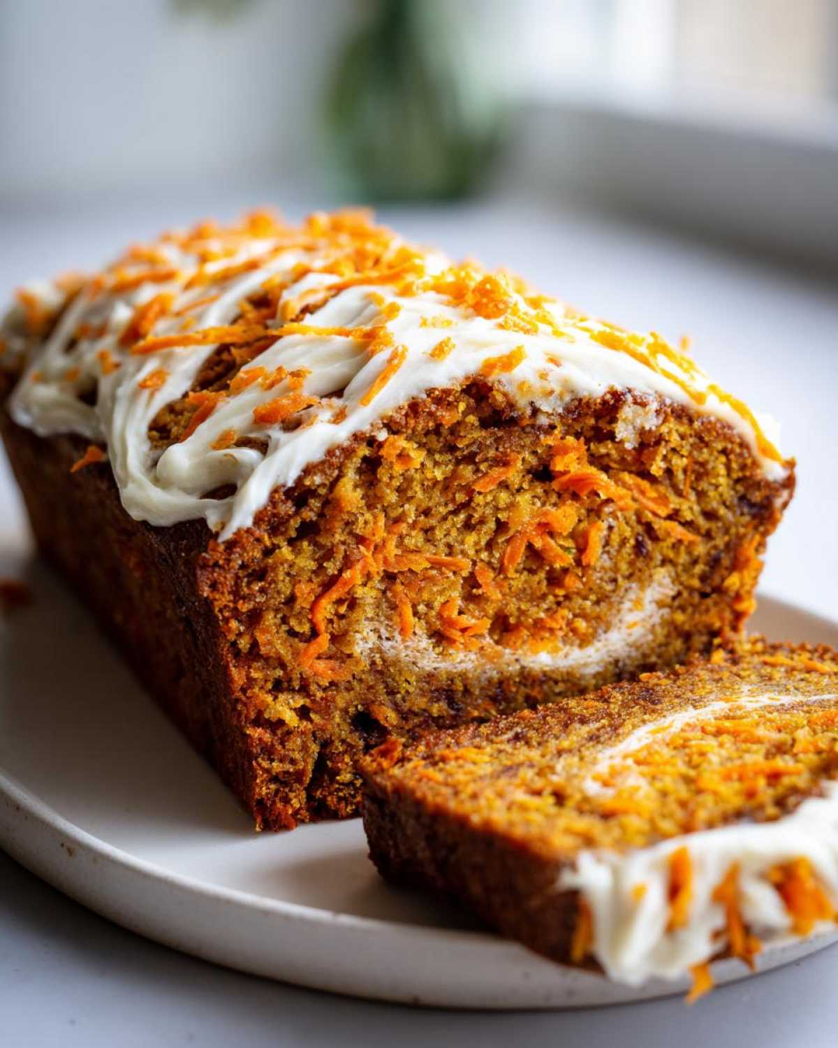 A loaf of carrot cake banana bread, sliced, topped with cream cheese frosting and shredded carrots.