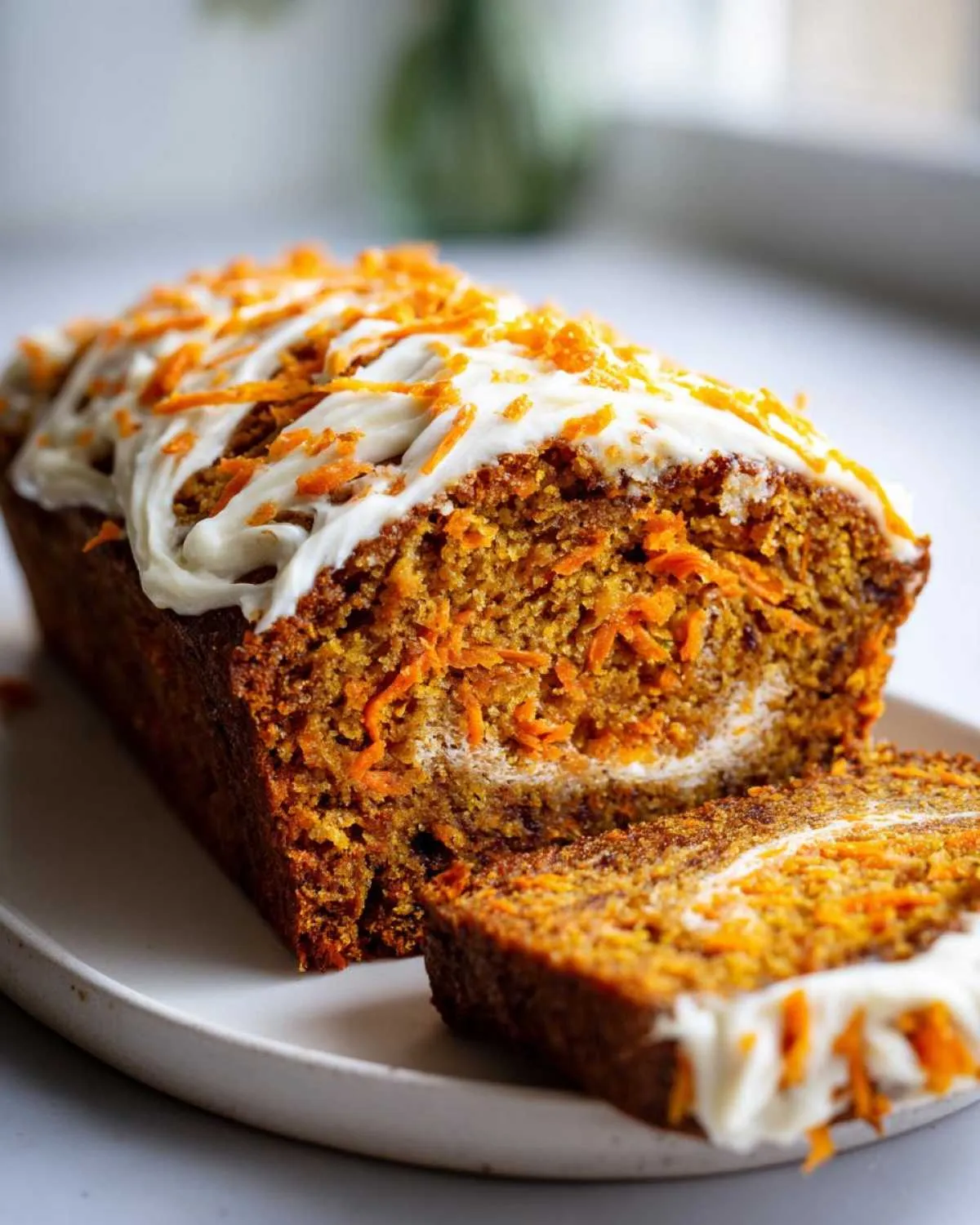 A loaf of carrot cake banana bread, sliced, topped with cream cheese frosting and shredded carrots.