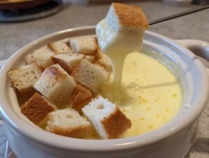 A piece of toasted bread is dipped into a white pot of creamy cheese fondue recipe, showing the melted cheese dripping.