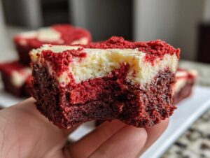 A hand holding a rich, fudgy red velvet brownie bite with a visible cheesecake swirl layer on top.
