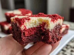 A hand holding a rich, fudgy red velvet brownie bite with a visible cheesecake swirl layer on top.