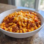 A close-up of a hearty bowl of ground beef goulash mixed with elbow macaroni and topped with melted cheddar cheese.
