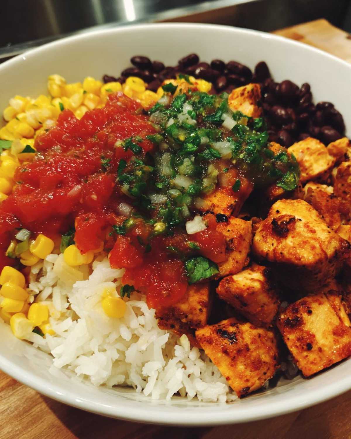 Close-up of a white bowl filled with ingredients for a chicken burrito bowl, including seasoned chicken, rice, corn, black beans, salsa, and pico de gallo.