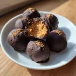 A plate of chocolate covered peanut butter balls, one cut open to reveal the creamy peanut butter center.