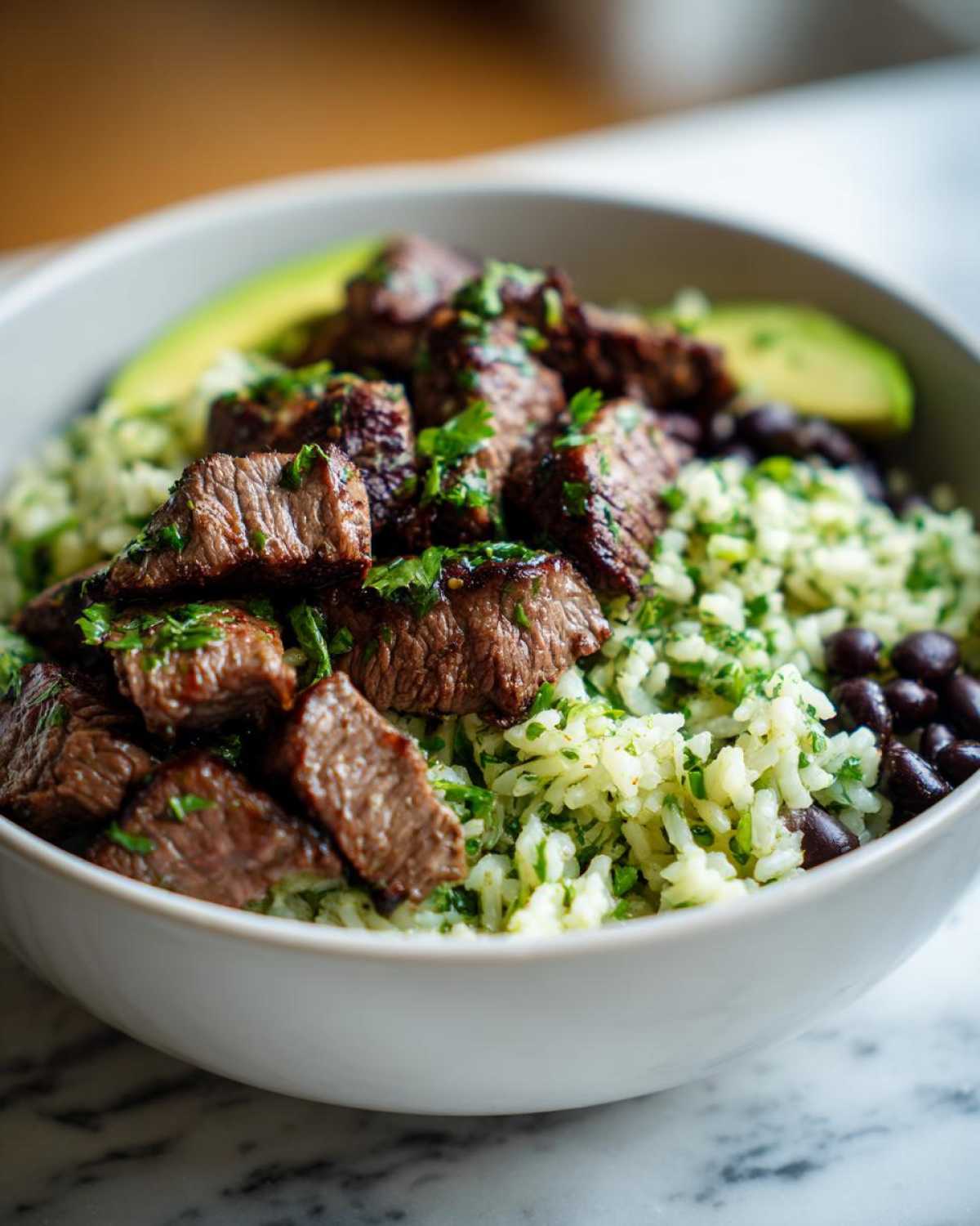 Close-up of a white bowl filled with cilantro lime steak bowls, featuring seasoned steak, cilantro rice, black beans, and avocado slices.