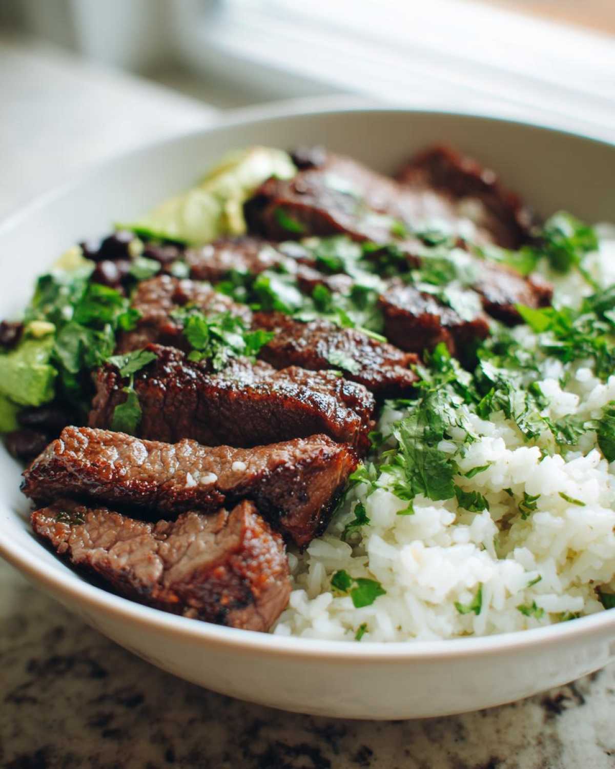 Close-up of sliced, marinated steak served over white rice, topped with fresh cilantro in a cilantro lime steak bowls preparation.