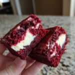 A hand holding two halves of a rich, red velvet brownie bite showing a creamy white cheesecake swirl center.