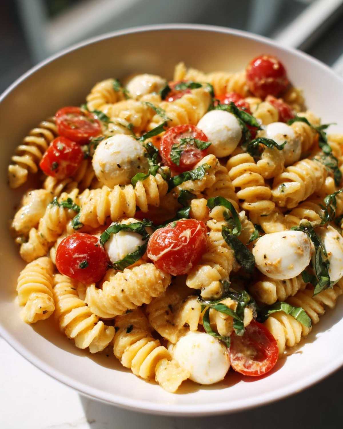 A close-up view of creamy caprese pasta salad featuring fusilli pasta, cherry tomatoes, mozzarella balls, and fresh basil.