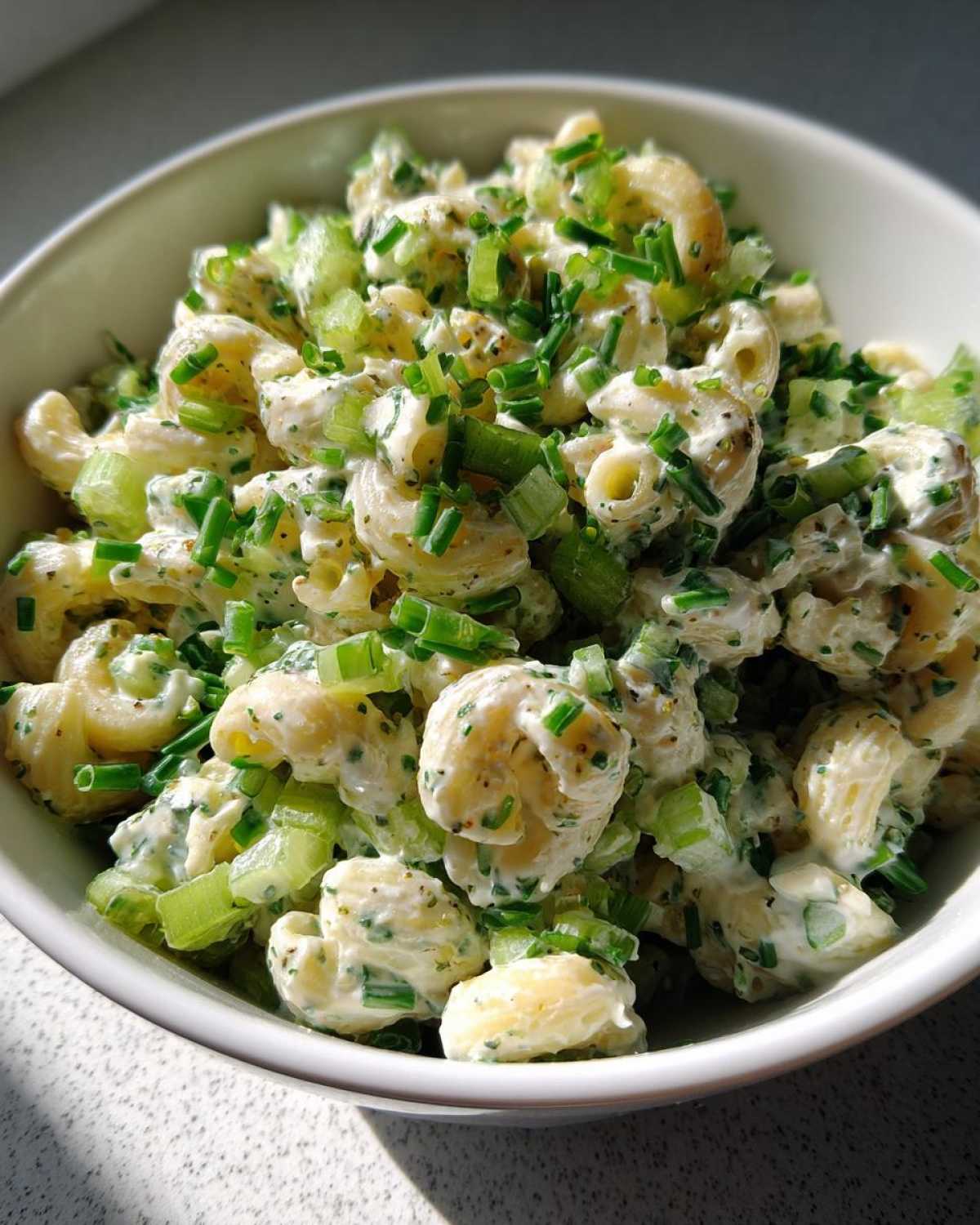 Close-up of a creamy cottage cheese pasta salad with elbow macaroni, chopped celery, and fresh chives in a white bowl.