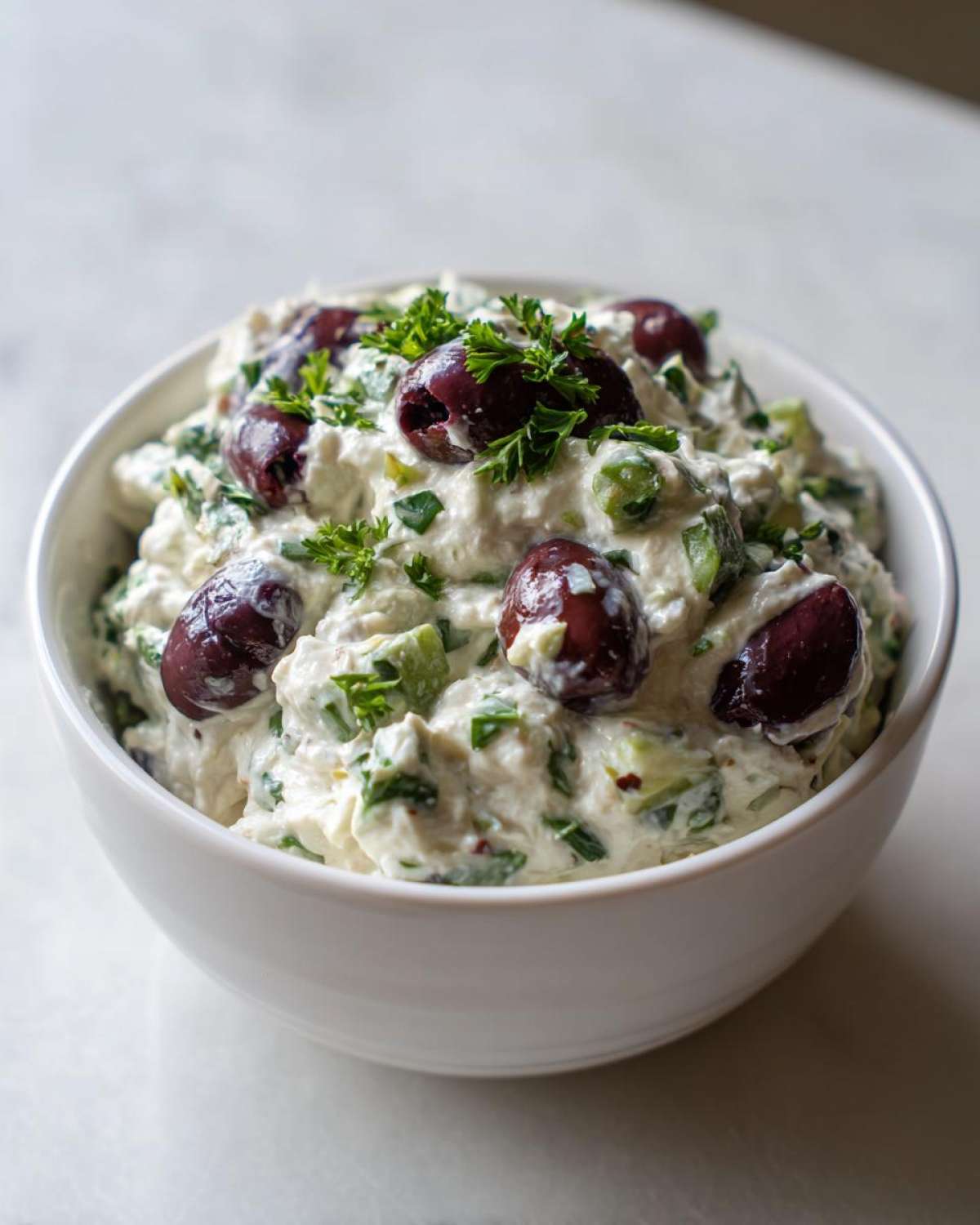 A creamy, white olive lovers dream dip topped with Kalamata olives and fresh parsley in a white bowl.