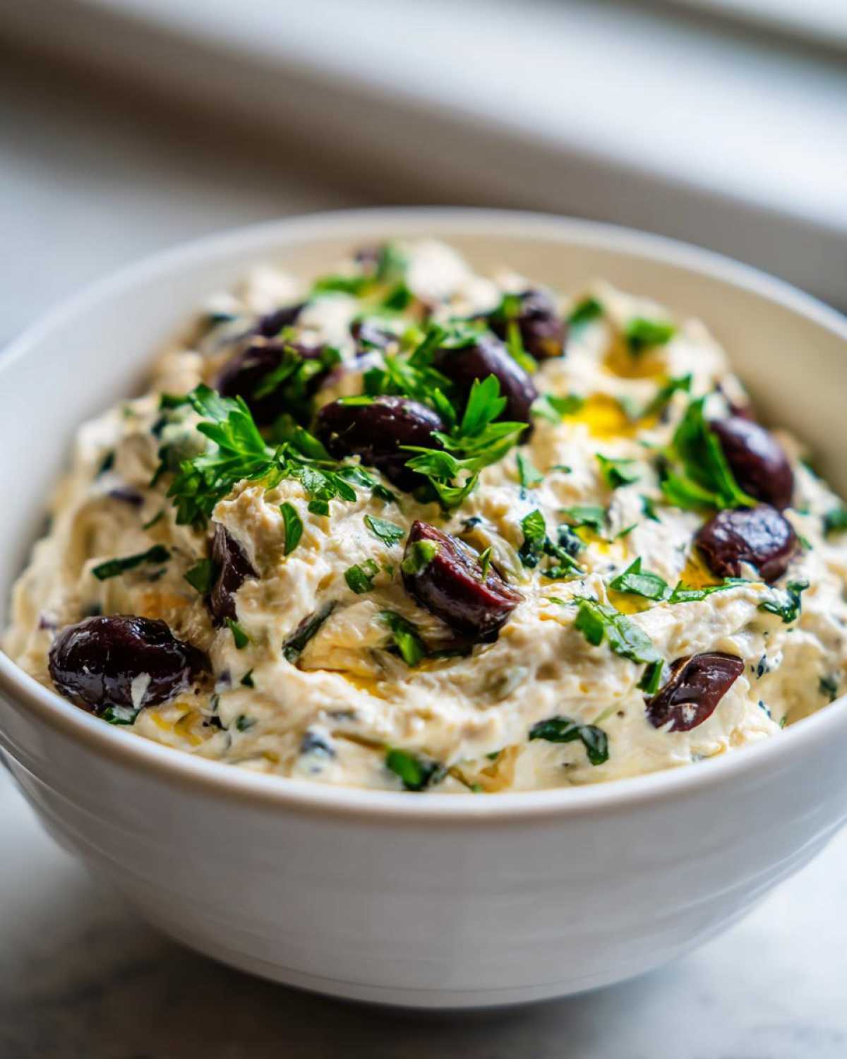 A creamy, light-colored olive lovers dream dip garnished with whole and halved Kalamata olives and fresh parsley.
