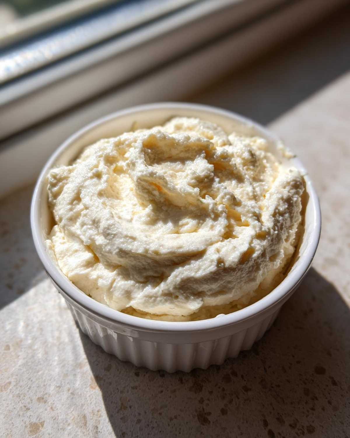 A small white ramekin filled with thick, creamy white pizza sauce, highlighted by sunlight.