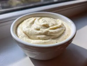 A close-up of thick, creamy white pizza sauce swirled in a small white ramekin, sitting on a windowsill.