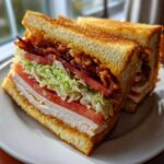 A perfectly cut, towering crispy ultimate club sandwich showing layers of toasted bread, turkey, tomato, lettuce, and bacon.