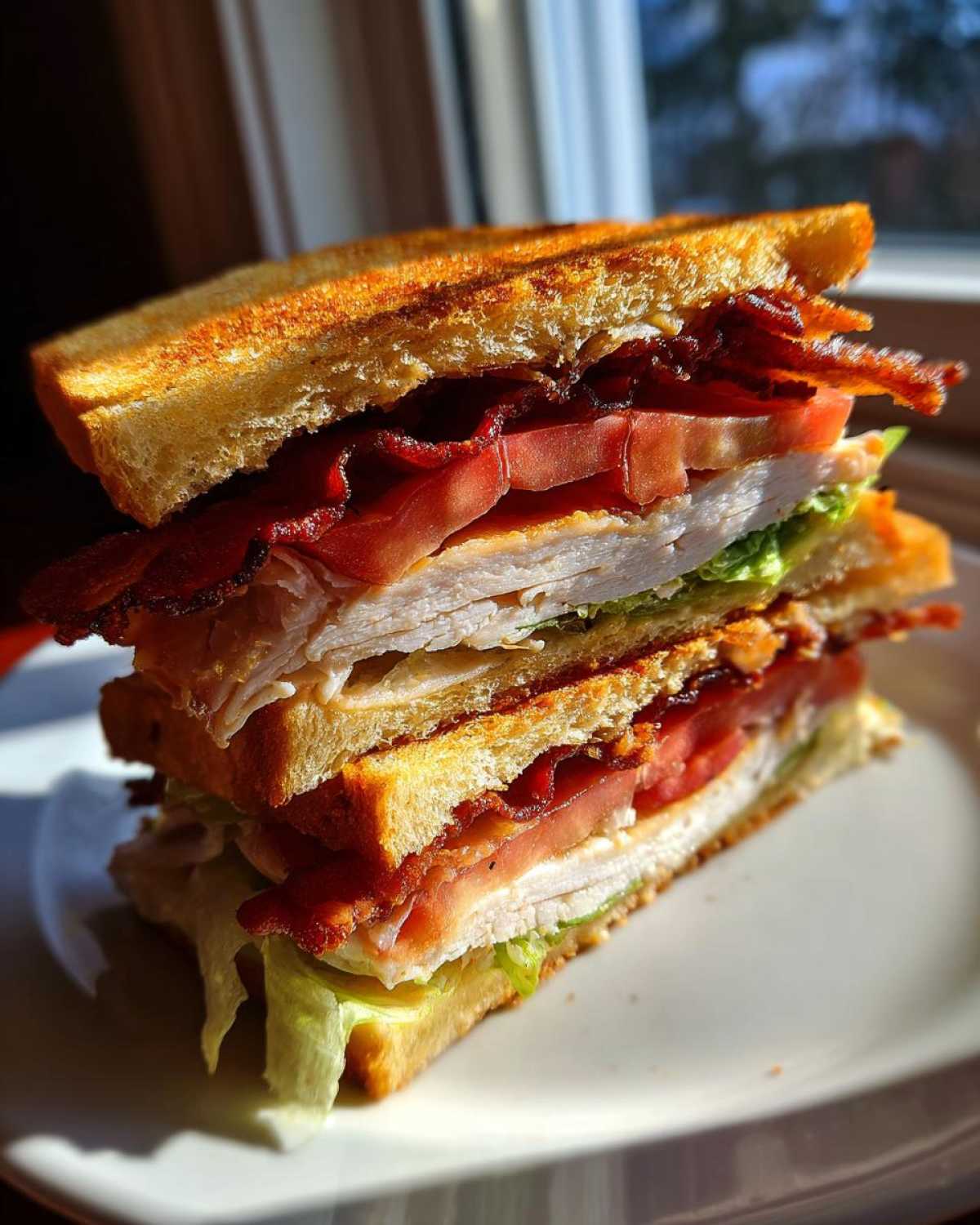 A tall, double-decker crispy ultimate club sandwich featuring toasted bread, turkey, bacon, tomato, and lettuce, served on a white plate.